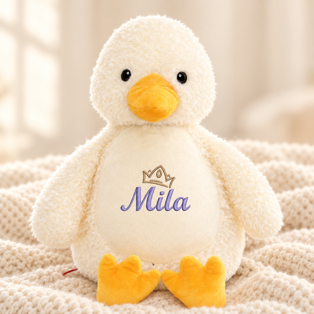 Personalisiertes Kuscheltier Ente mit Name & Krone – Beispiel Mila auf cremefarbener Decke